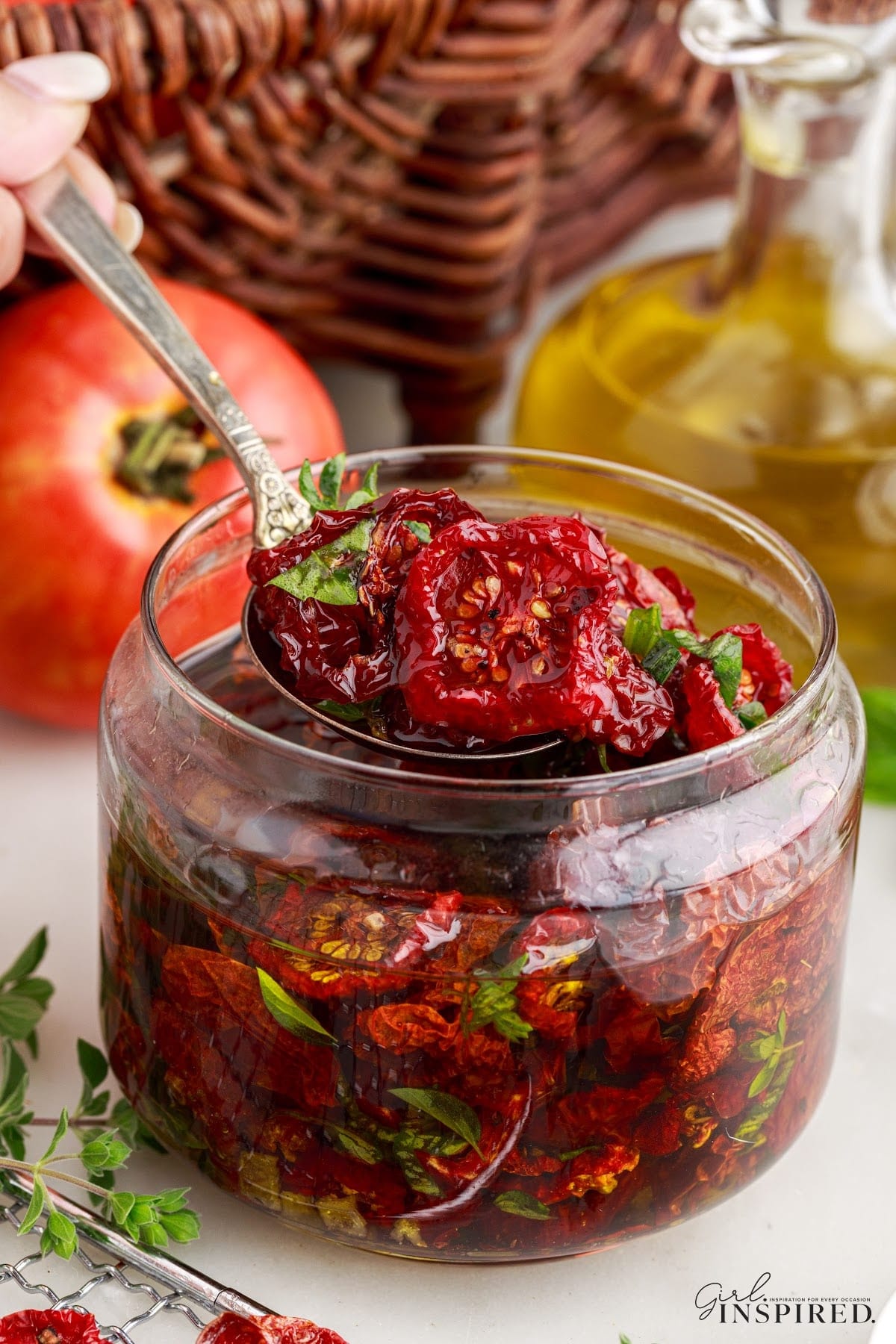 Sun-dried tomatoes in a jar packed in oil with a spoon. A jar of olive oil and fresh whole tomatoes in the background.