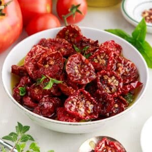 Bowl of sun-dried tomatoes with fresh whole tomatoes and herbs in the background.