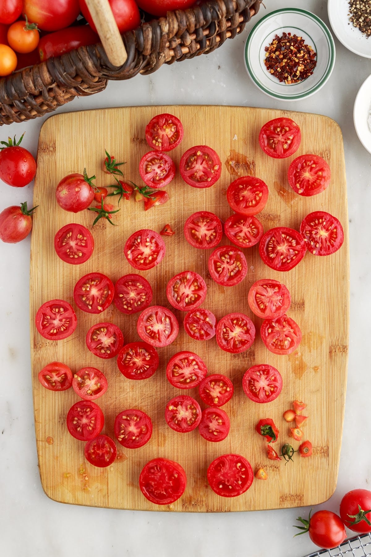 Sliced fresh tomatoes on a wooden kitchen board with seasonings in small bowls and a basket of fresh whole tomatoes in the background.